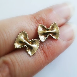 May include: A pair of gold-colored earrings shaped like pasta. The earrings are in the shape of a bow tie pasta, also known as farfalle.