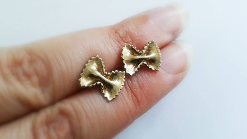 May include: A pair of gold-colored earrings shaped like pasta. The earrings are in the shape of a bow tie pasta, also known as farfalle.