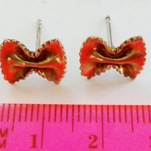 May include: A pair of gold-colored earrings shaped like pasta, with a red-orange glaze. The earrings are about 1 cm long and are shown next to a ruler.