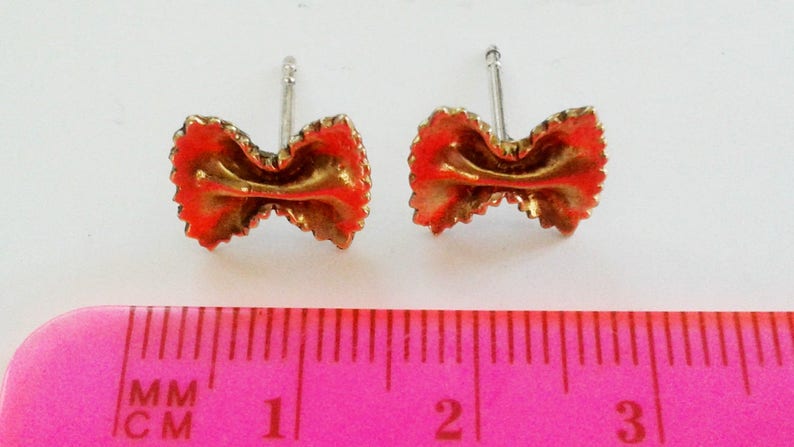 May include: A pair of gold-colored earrings shaped like pasta, with a red-orange glaze. The earrings are about 1 cm long and are shown next to a ruler.