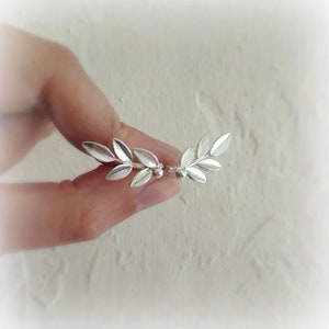 Könnte beinhalten: Silberne Blatt-Ohrclips mit einem zarten Design. Die Ohrclips sind in Form eines Zweigs mit kleinen Blättern und einer kleinen Perle am Ende gestaltet.