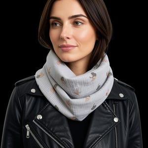 May include: A light gray infinity scarf featuring a repeating pattern of small, muted-colored ginkgo leaf shapes. The scarf is made of a textured, crinkled fabric and is worn around the neck over a black leather jacket with silver metal zippers.