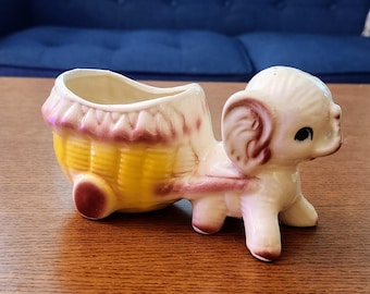 May include: A vintage ceramic planter shaped like an elephant pulling a yellow and pink wagon. The elephant is cream-colored with brown accents. The wagon has a basket-like design.