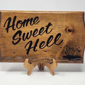 May include: Wooden sign with the phrase "Home Sweet Hell" in black script. The sign has a natural wood grain finish and is supported by a wooden easel. Decorative elements include birds and cattails.
