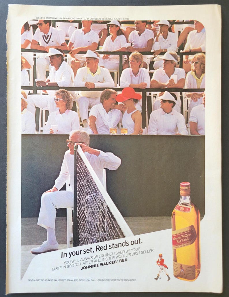 May include: An advertisement for Johnnie Walker Red Label whisky, featuring a tennis match scene with spectators in white. The image includes a bottle of whisky and the text "In your set, Red stands out."