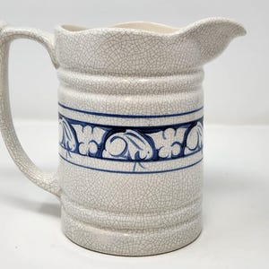 May include: Cream-colored ceramic pitcher with a blue floral design band. The pitcher has a large handle and a spout for pouring. The surface has a crackle glaze effect, adding to its vintage aesthetic. A decorative piece for serving beverages.