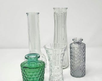 May include: An assortment of glass vases in various shapes and sizes. The vases are clear, green, and gray, with textured and smooth surfaces. The arrangement includes tall, slender vases and shorter, decorative ones.