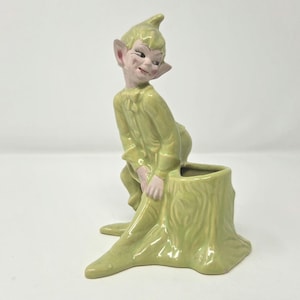 May include: A glazed ceramic figurine of a pointed-ear elf with a light green complexion and matching clothing, leaning against a hollow tree stump. The elf has a mischievous expression, and the hollow stump serves as a small planter or vessel.