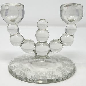 May include: Clear glass candle holder with two candle cups and a central decorative element. The base is round with a detailed pattern. The candle holder is made of clear glass.