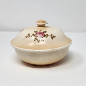 May include: Cream-colored ceramic dish with a lid, featuring a pink rose floral design. The lid has a small knob and the dish has a slightly aged appearance. A decorative piece for the home.
