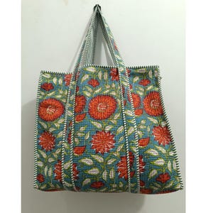 Op de afbeelding: Een grote draagtas met een bloemenpatroon. De tas heeft een turquoise achtergrond met grote rode bloemen en groene bladeren. De handvatten en randen zijn afgewerkt met een streepjespatroon. De tas hangt tegen een witte muur.