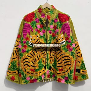 May include: An olive green velvet jacket featuring vibrant, multi-coloured floral embroidery and two large tiger motifs on the front panels. The garment includes a collar, long sleeves, and the text CraftofIndianShop printed across the centre.