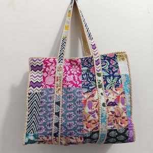 Könnte beinhalten: Eine Patchwork-Tragetasche mit einer Vielzahl von Blumen- und geometrischen Mustern in den Farbtönen Rosa, Blau, Lila und Weiß. Die Tasche hat lange Griffe und eine beige Einfassung. Die Tasche hängt an einer weißen Wand.