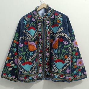 Op de afbeelding: Donkerblauwe jas met kleurrijke bloemenborduursels. De jas heeft een opstaande kraag, een ritssluiting en een gestreepte voering. De borduursels bevatten bloemen in rood, oranje, blauw, paars en groen.