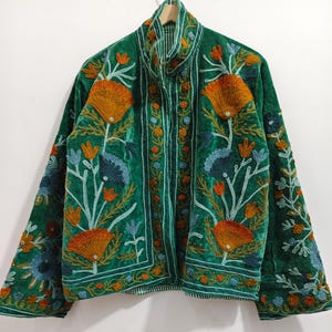 May include: A green velvet jacket with intricate floral embroidery in orange, blue, and cream. The jacket features a high collar and button-up front. The sleeves are long and the embroidery covers the front, back, and sleeves.