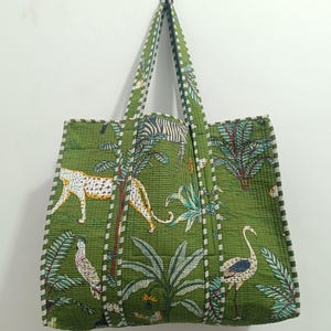 Könnte beinhalten: Grüne Tragetasche mit Dschungelmotiv mit Zebras, Leoparden und Vögeln. Die Tasche hat eine gesteppte Textur und weiße Verzierungen entlang der Kanten und Träger. Die Tragetasche hängt an einer weißen Wand.