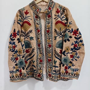 May include: A beige jacket featuring a mandarin collar and long sleeves, decorated with intricate floral embroidery in shades of red, blue, and yellow. The garment is open at the front and displays a textured fabric surface throughout the design.