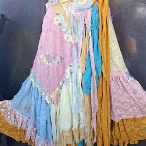 Op de afbeelding: Een kleurrijke, bohemien rok met een patchwork ontwerp. De rok heeft verschillende kanten stoffen in roze, blauw, geel en crème. De rok is versierd met bloemversieringen en lange, vloeiende linten.