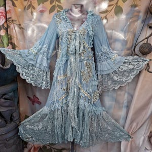 May include: A light blue dress featuring long, flared bell sleeves with layered ruffles and intricate lace trim. The garment includes a V-neckline with a tie closure, decorative floral embroidery, and a textured, layered skirt with lace panels.