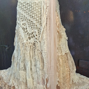 Puede incluir: Falda crema en capas con intrincados detalles de encaje y ganchillo. La falda presenta múltiples texturas, incluyendo flecos y tul, creando una estética bohemia. Un panel de tela color rubor añade un suave contraste al diseño.