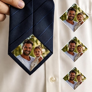 Puede incluir: Corbata azul marino con un patrón de rayas diagonales. La corbata presenta una foto en forma de diamante de un hombre y un niño. Tres fotos adicionales están prendidas a una camisa blanca.