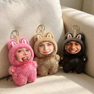 May include: Three plush keychains featuring fuzzy animal hoods in pink, tan, and dark brown. Each keychain has a metal ring and a cutout in the hood displaying a realistic human face. The plush figures have small ears and soft fur textures.