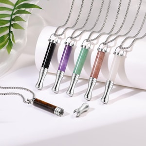 Puede incluir: Una colección de collares con colgantes de piedras preciosas y cadenas plateadas. Los colgantes cilíndricos están disponibles en varios colores, como negro, morado, verde, marrón y blanco. Un colgante está separado, mostrando un pequeño compartimento.