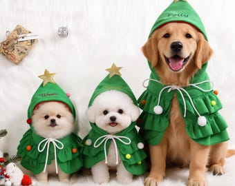 May include: Three dogs wearing green hooded capes designed to resemble Christmas trees. Each cape features a gold star at the top, white drawstring ties, and colorful pom-poms. The names Winnie, Sami, and Polly are embroidered on the hoods.