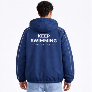 Könnte beinhalten: Marineblaue Kapuzenjacke mit weißer Paspelierung und dem weißen Aufdruck "KEEP SWIMMING" auf dem Rücken. Die Jacke hat eine Kapuze mit Kordelzug und einen Reißverschluss. Der Text befindet sich über einem Wellenmuster.