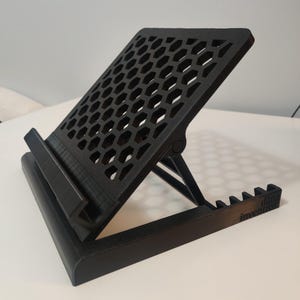 May include: A black, adjustable book stand with a honeycomb pattern on the angled surface. The stand has a base and a ledge to hold the book in place. The text "dani invent" is printed on the bottom edge.