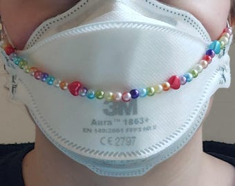 May include: A white face mask with a colorful beaded chain. The chain features a variety of beads in colors like red, blue, green, and yellow. The mask has the text "3M Aura 1863+" printed on it.