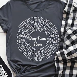 A gray t-shirt with a white circular design, a pair of black and white sneakers, and a black and white plaid shirt are arranged on a white surface.