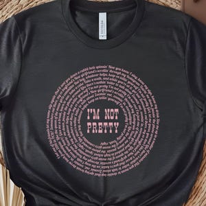 May include: Dark gray t-shirt with a circular design in pink text that reads "I'M NOT PRETTY" in the center. The design is surrounded by a spiral of smaller text. The shirt is knotted at the bottom.