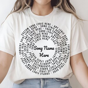 A woman wearing a white t-shirt with a circular design and the text "Song Name Here" printed on it, sitting cross-legged on the floor.