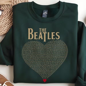 May include: Dark green sweatshirt featuring "The Beatles" in gold lettering above a heart shape filled with song lyrics. A small red heart is at the bottom of the heart shape. The sweatshirt is folded, with a pair of jeans and shoes in the background.