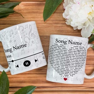 May include: Two white ceramic mugs with musical note designs and the text "Song Name" and "Artist". One mug features a music player interface. The other mug has song lyrics and a red heart. The mugs are on a wooden surface.