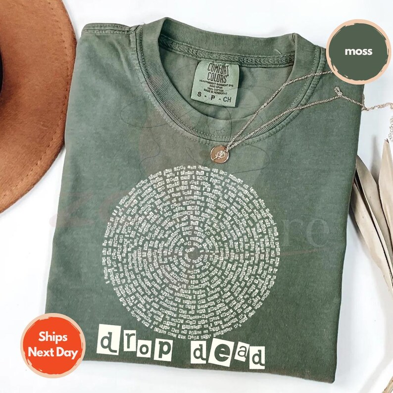 May include: A moss green t-shirt with a circular design of text and the words "drop dead" in white block letters. A silver necklace with a small pendant rests on the shirt. The shirt is a Comfort Colors brand.