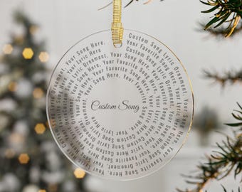 May include: A clear, round ornament with gold trim and a gold ribbon for hanging. The ornament features the text "Custom Song" in the center, surrounded by a circular pattern of text. The text reads "Your Favorite Song Lyric Ornament."