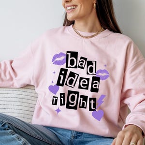 May include: A light pink crewneck sweatshirt featuring the black text "bad idea right" in a ransom-note style font. The design is accented with purple lip prints, purple hearts, a purple lightning bolt, and small four-pointed star shapes.
