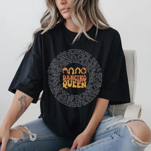 A woman wearing a black t-shirt with a graphic design and text, sitting on a chair.