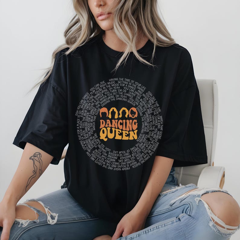 A woman wearing a black t-shirt with a graphic design and text, sitting on a chair.