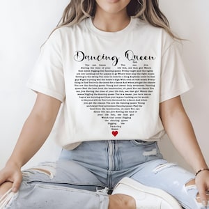 May include: Cream-colored t-shirt featuring the words "Dancing Queen" in a stylized font. The lyrics of the song are printed in a heart shape below the title, with a small red heart at the bottom. A casual, everyday wear tee.