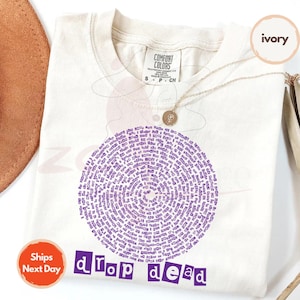 May include: Ivory-colored t-shirt with a circular purple design composed of text. The words "drop dead" are printed in purple block letters below the design. A gold necklace and a brown hat are visible.