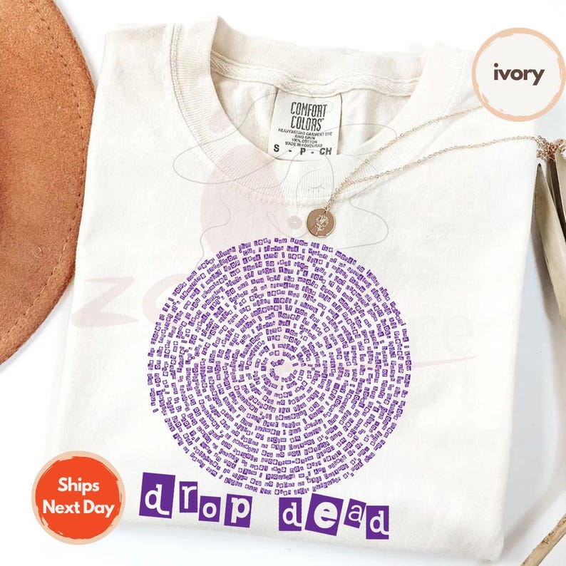 May include: Ivory-colored t-shirt with a circular purple design composed of text. The words "drop dead" are printed in purple block letters below the design. A gold necklace and a brown hat are visible.