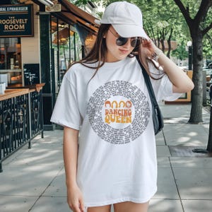 A woman wearing a white t-shirt with a graphic design and a cap is standing on a sidewalk.