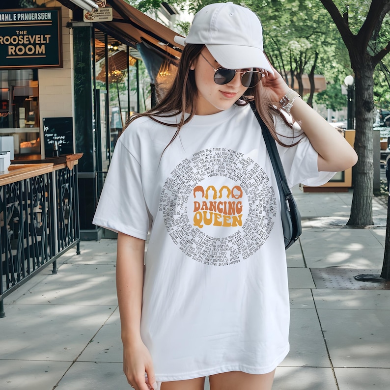 A woman wearing a white t-shirt with a graphic design and a cap is standing on a sidewalk.