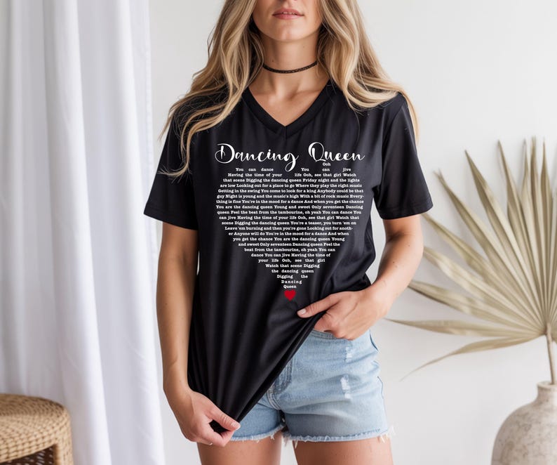 A woman wearing a black t-shirt with white text and a red heart design stands in front of a white curtain and a palm plant.