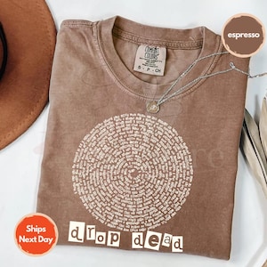 May include: Espresso-colored t-shirt with a circular design of text and the words "drop dead" in a bold, white font. The shirt has a crew neck and short sleeves. A silver necklace with a pendant is draped across the shirt.