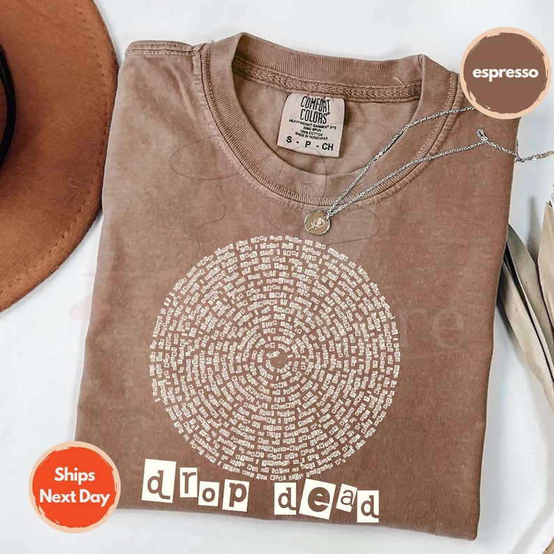 May include: Espresso-colored t-shirt with a circular design of text and the words "drop dead" in a bold, white font. The shirt has a crew neck and short sleeves. A silver necklace with a pendant is draped across the shirt.