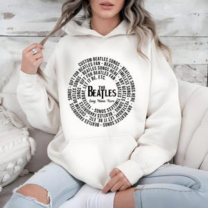 May include: Cream-colored hoodie featuring a circular design with the text "The Beatles" and song titles. The design includes phrases like "Custom Beatles Songs" and "Gift for Beatles Fan." The hoodie is a comfortable, casual style.